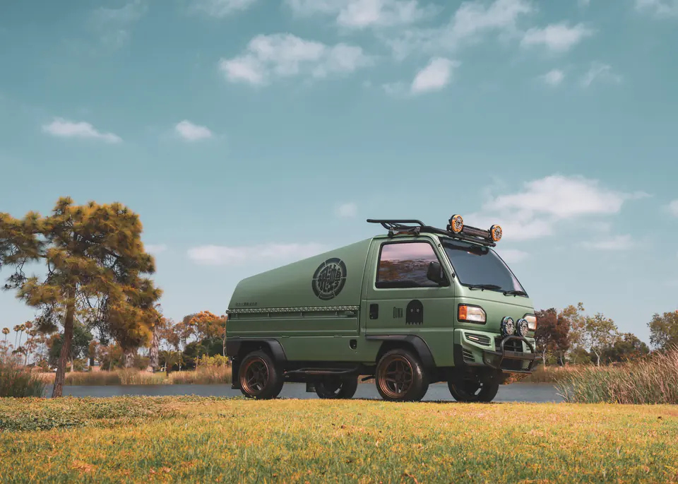 Green Honda Acty Kei Truck camper conversion with a roof rack and LED driving lights beside a lake