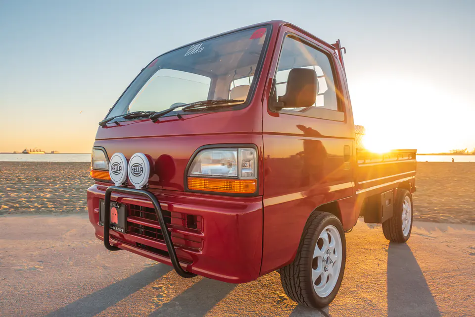 Red Honda Acty Kei Truck with a black bull bar and round driving lights parked on a California beach at sunset